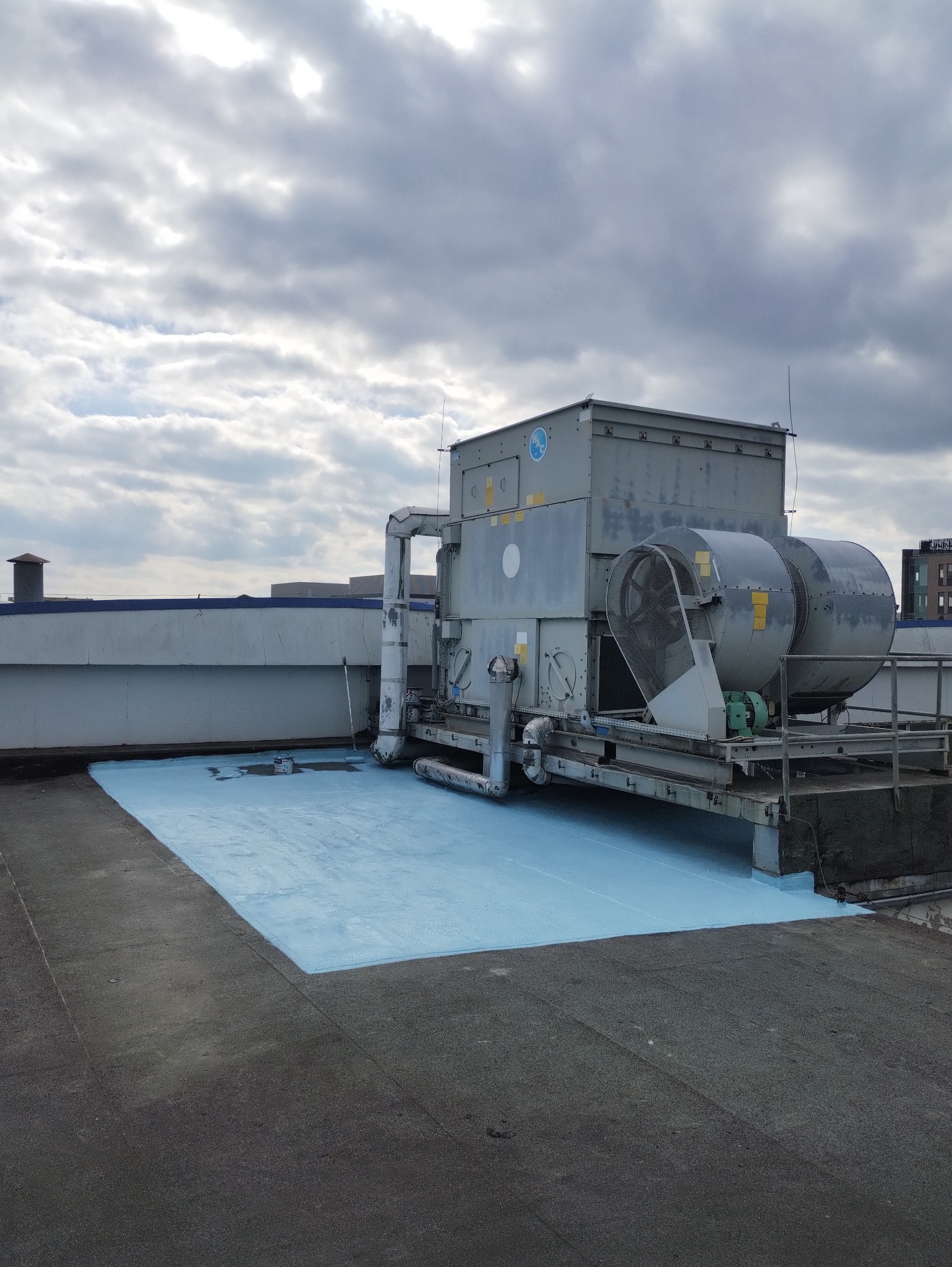 Płaski dach pokryty świeżą, jasnoniebieską warstwą hydroizolacyjną, z widoczną instalacją wentylacyjną i fragmentami innych budynków w tle.