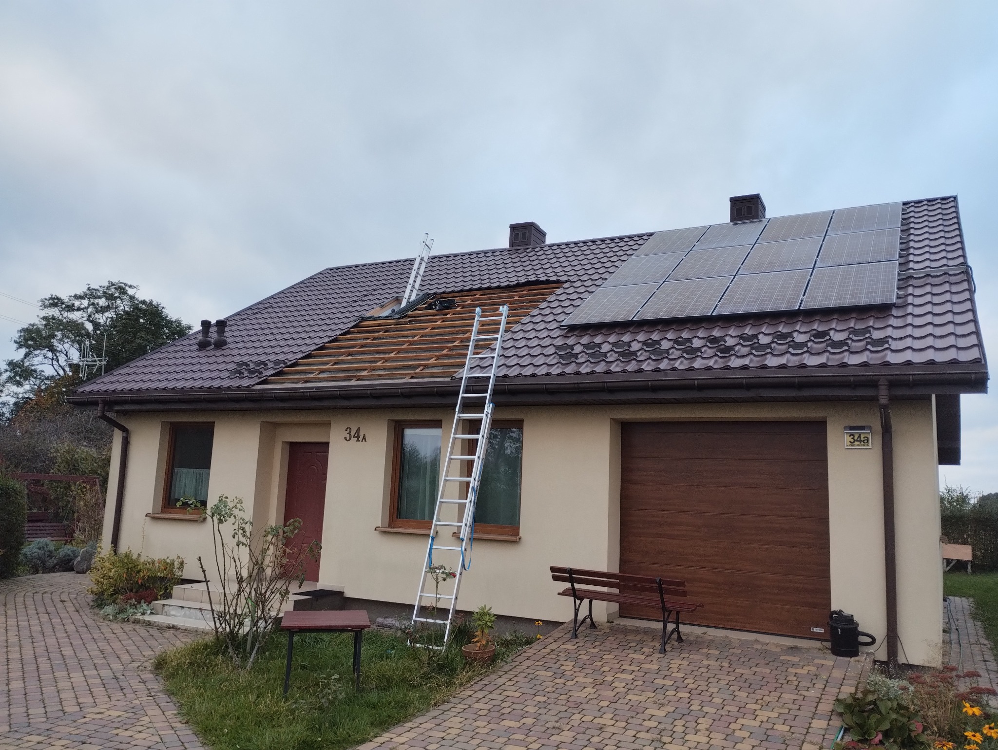 Dom jednorodzinny z brązowym dachem częściowo zdjętym, widoczne drewniane łaty, panele solarne i dwie drabiny oparta o dach.