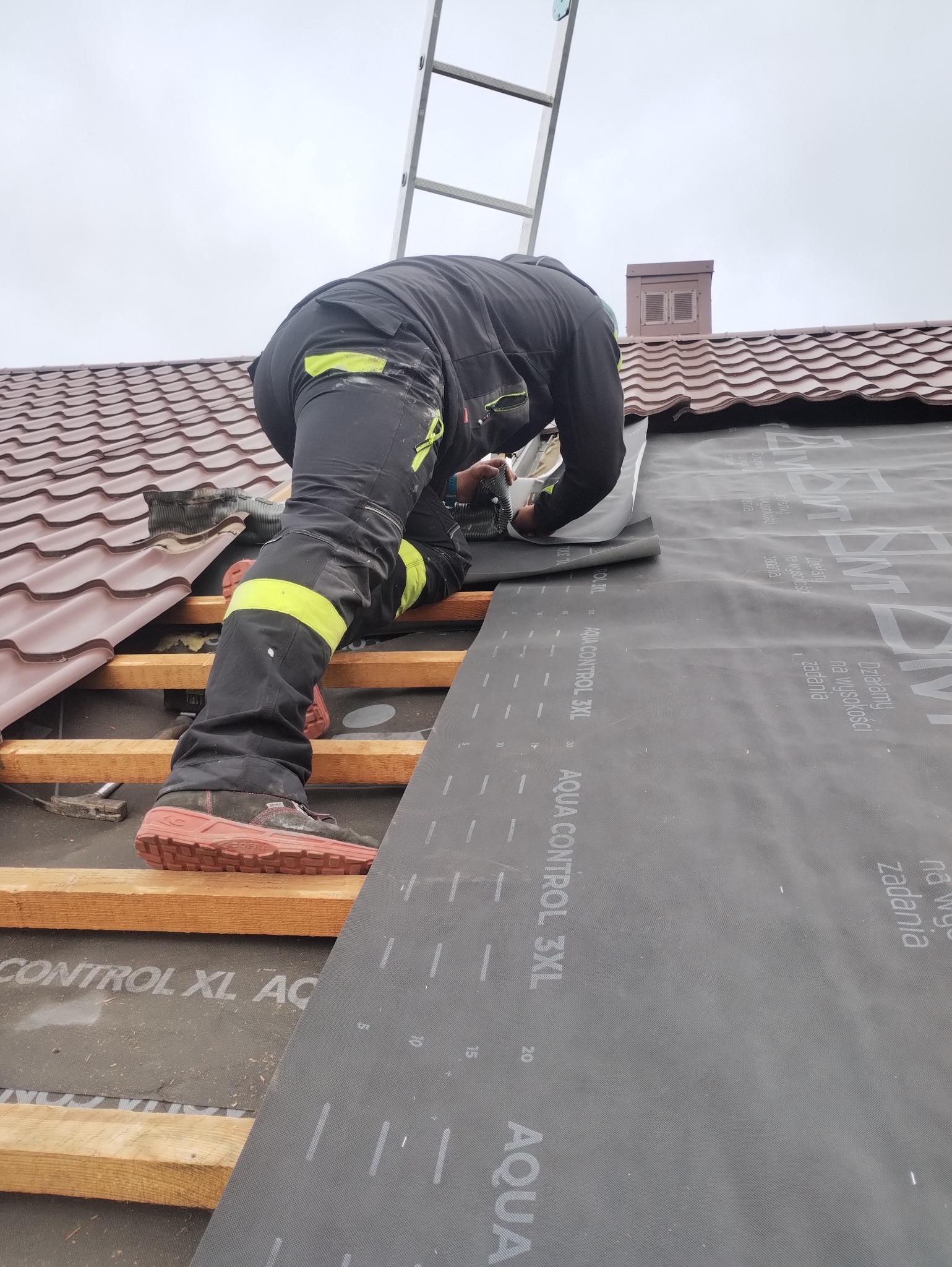 Pracownik w kombinezonie z odblaskowymi pasami układa membranę dachową Aqua Control 3XL na drewnianej konstrukcji dachu z brązową dachówką, widoczna drabina i komin.