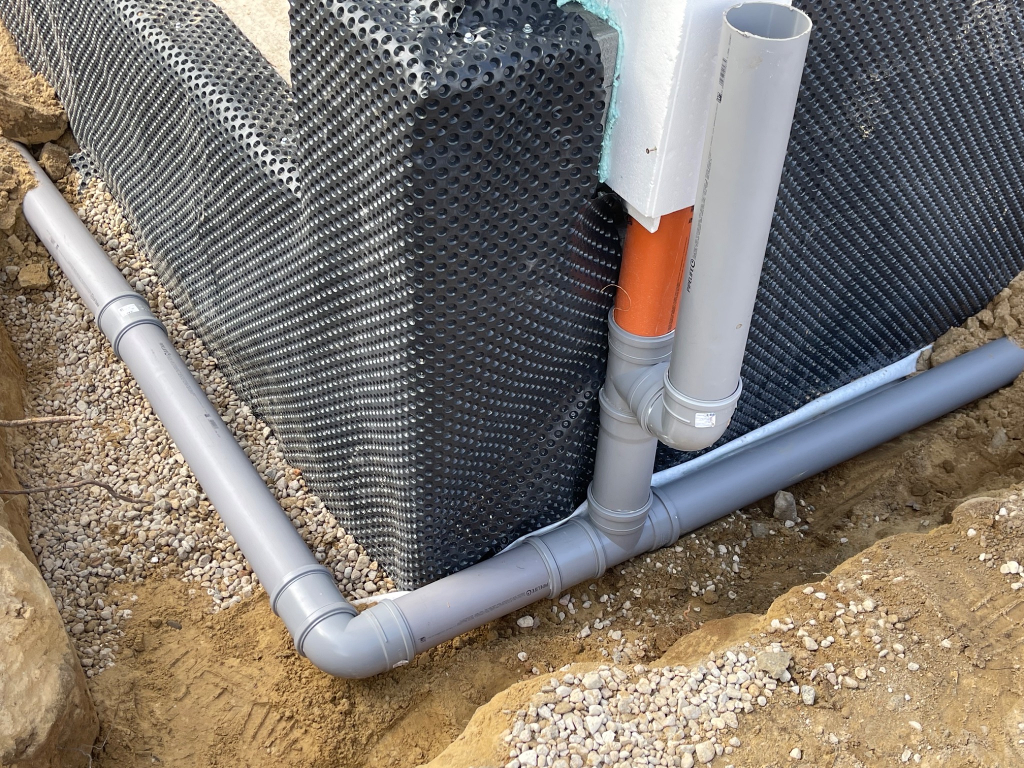 Fundament zabezpieczony folią kubełkową z odprowadzeniem wody rurami PCV w wykopie, obsypany żwirem. Widoczne łączenia rur i izolacja termiczna.