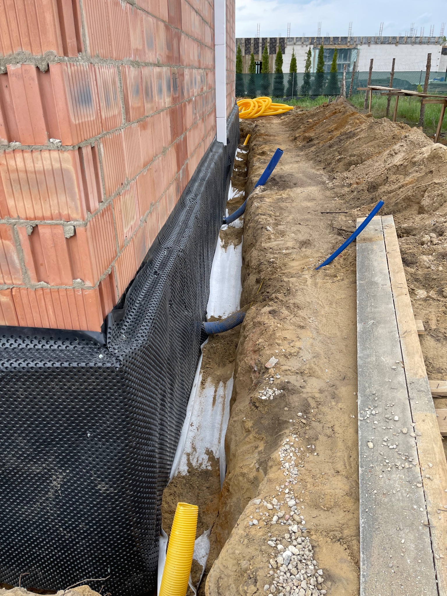 Fundament budynku w trakcie prac: izolacja pionowa z folii kubełkowej, drenaż opaskowy z rur drenarskich w wykopie. Widoczne materiały budowlane i narzędzia.