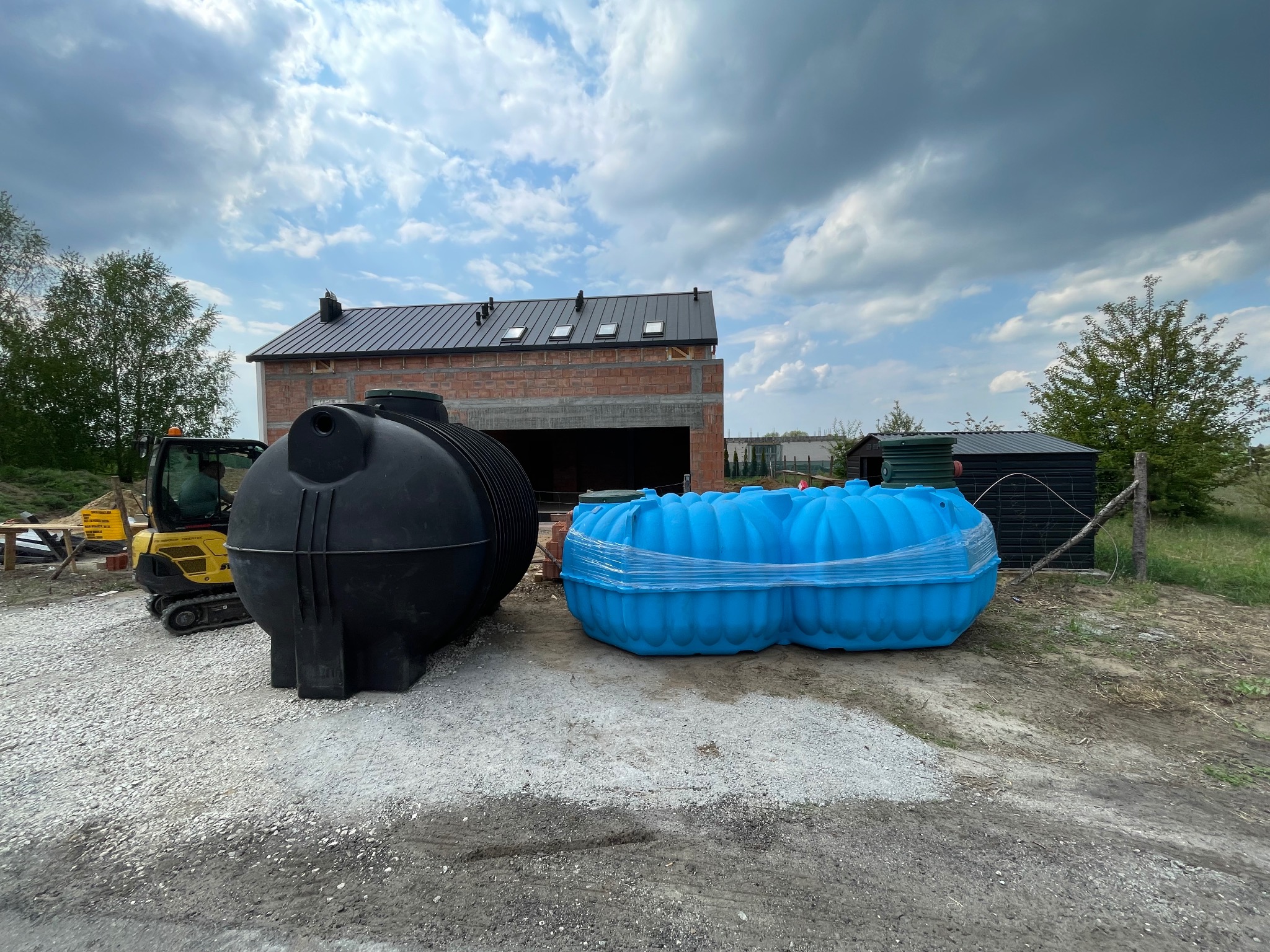 Szamba betonowe: Czarny i niebieski zbiornik na żwirowym podłożu, w tle budynek w trakcie budowy i minikoparka. Pochmurne niebo.