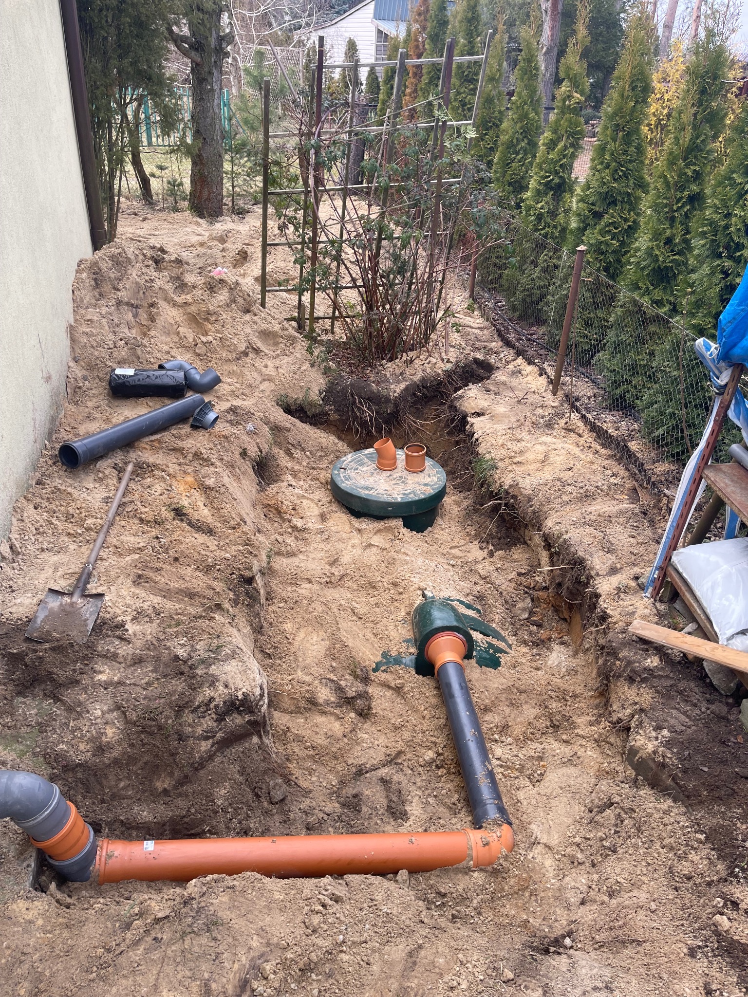 Montaż instalacji sanitarnej w wykopie ziemnym: rury kanalizacyjne, studzienka rewizyjna z pomarańczowymi rurami. W tle iglaki i drabina.
