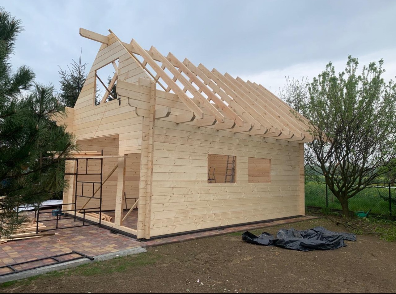 Surowy drewniany szkielet domu w trakcie budowy. Widoczne krokwie dachowe, ściany z bali i rusztowania. Budynek otoczony zielenią, w tle pochmurne niebo.