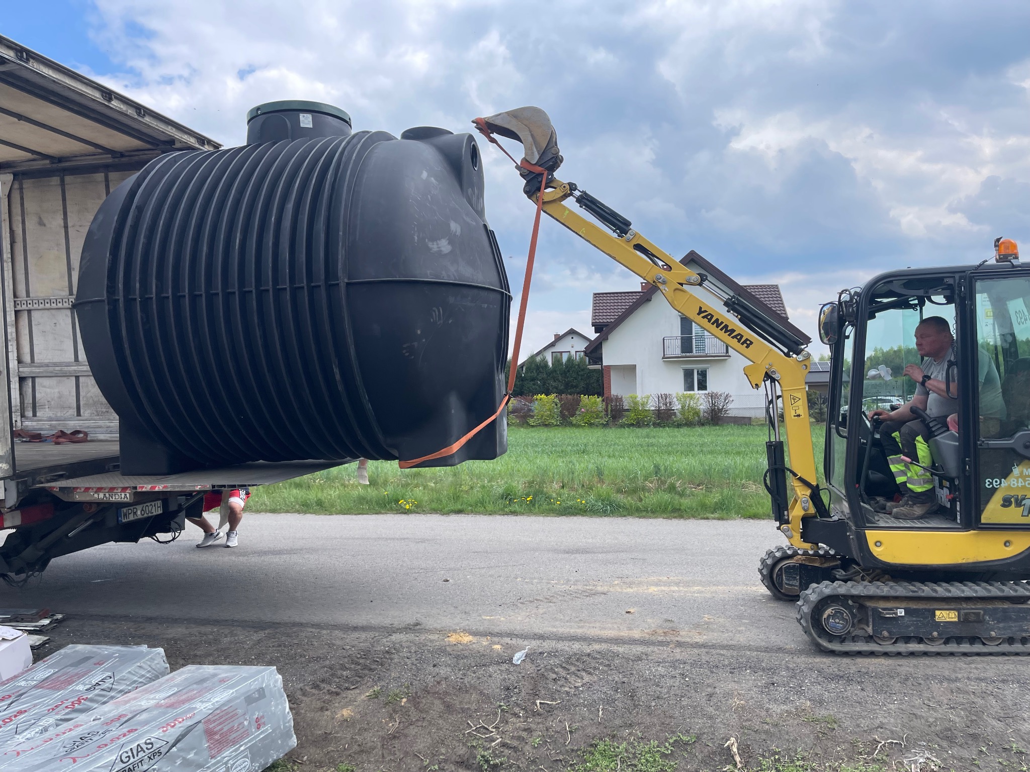 Żółta koparka Yanmar ostrożnie zdejmuje czarny zbiornik z naczepy ciężarówki na tle domów i zieleni. Widoczne pakiety materiałów izolacyjnych.