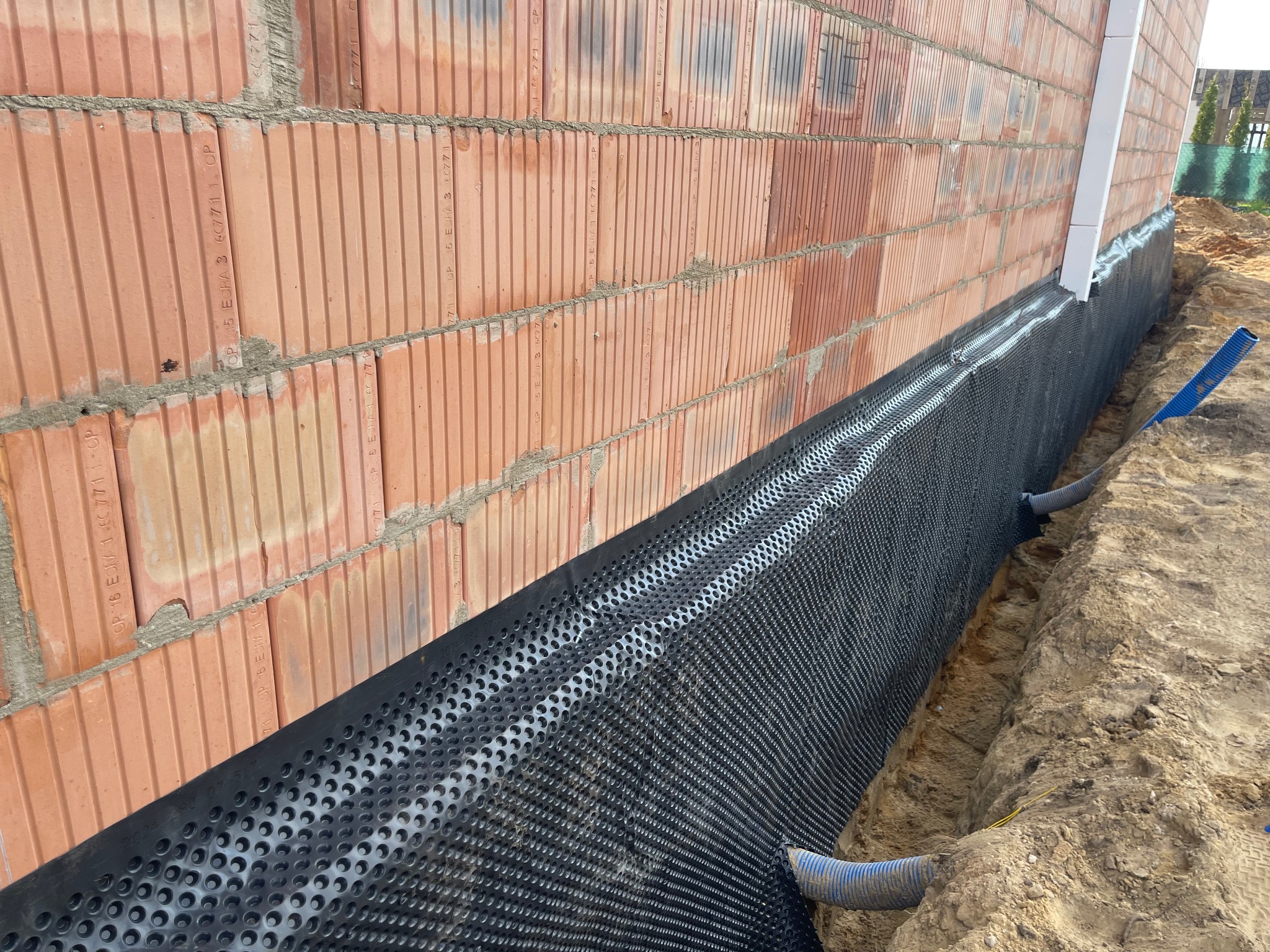 Ocieplenie fundamentów budynku z bloczków ceramicznych, zabezpieczone folią kubełkową i drenażem. Widok na fragment ściany i wykop z instalacją.
