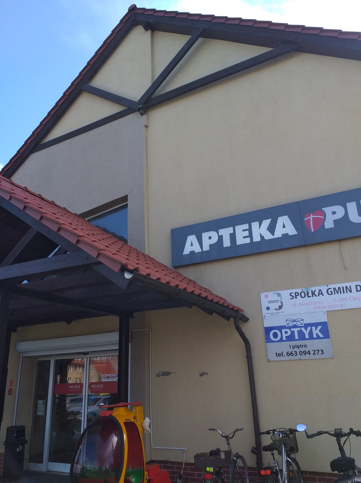 Budynek apteki z widoczną elewacją, dachem z dachówki i rynnami. Na ścianie szyldy reklamowe i informacyjne. Przed wejściem automat dla dzieci i zaparkowane rowery.