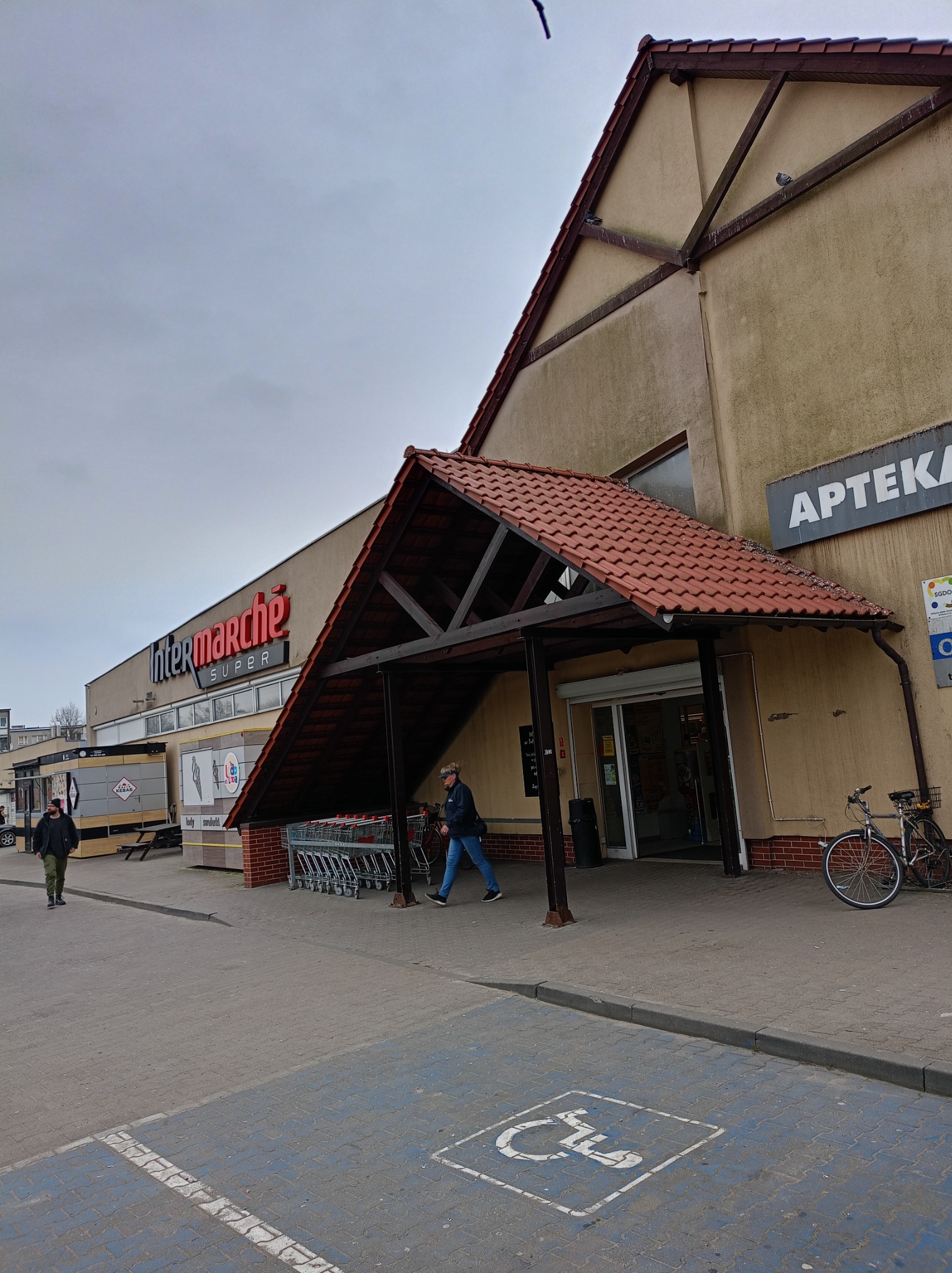 Fasada budynku Intermarche z wejściem zadaszonym czerwoną dachówką, widoczne oznakowanie apteki na ścianie, zaparkowany rower i osoby przechodzące obok.