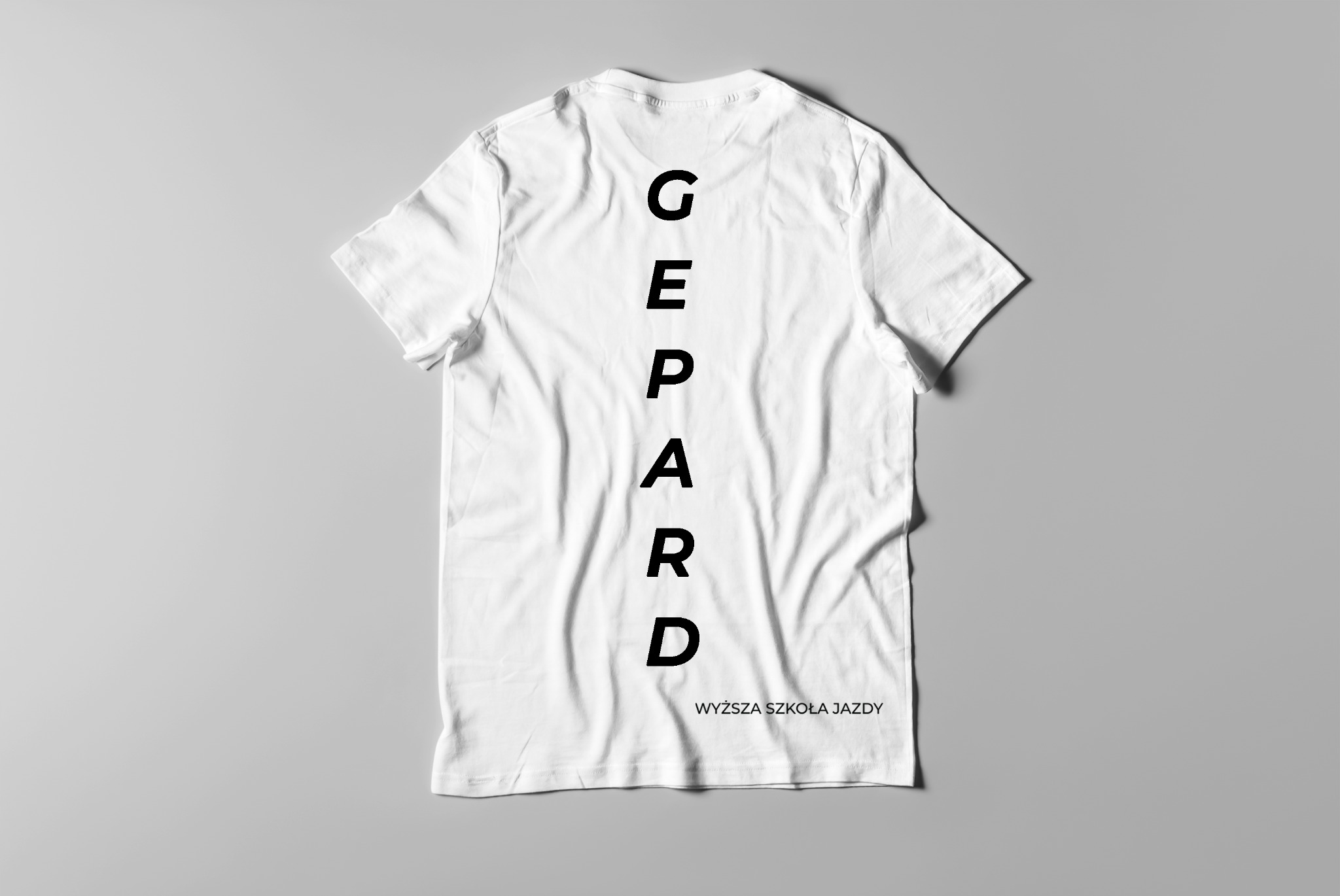 Biała koszulka z czarnym pionowym napisem 'GEPARD' i 'WYŻSZA SZKOŁA JAZDY' poniżej, leżąca na szarym tle.