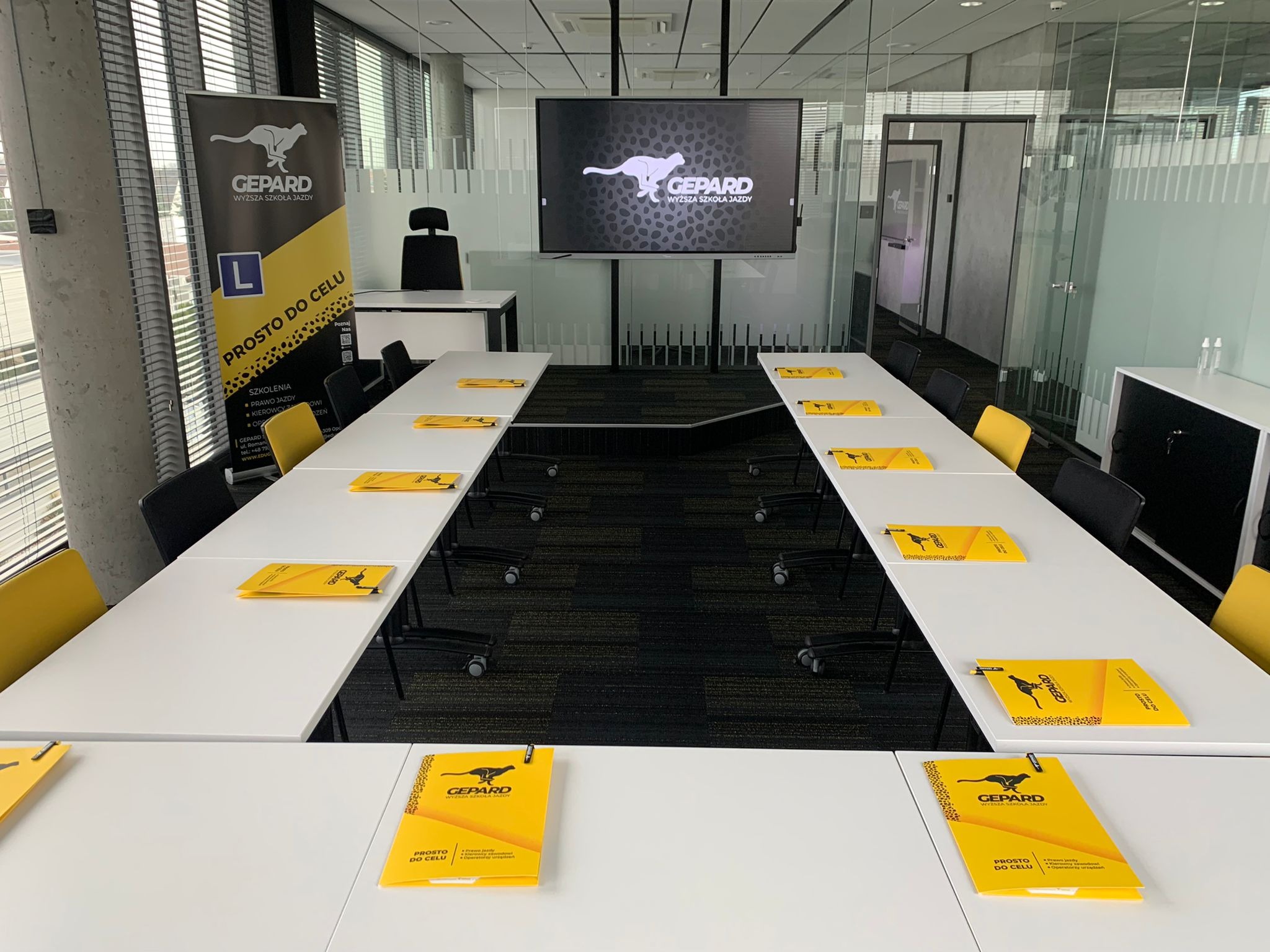 Sala szkoleniowa z białymi stołami, czarnymi i żółtymi krzesłami, oraz materiałami informacyjnymi z logo geparda na stołach. Na ścianie widoczny ekran z logiem firmy. Baner reklamowy z logiem...