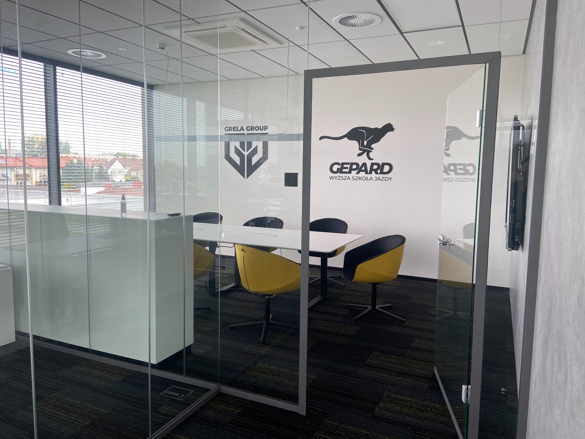 Nowoczesna sala konferencyjna za szklaną ścianą z widocznym logo 'Gepard Wyższa Szkoła Jazdy' oraz logo 'Grela Group'.