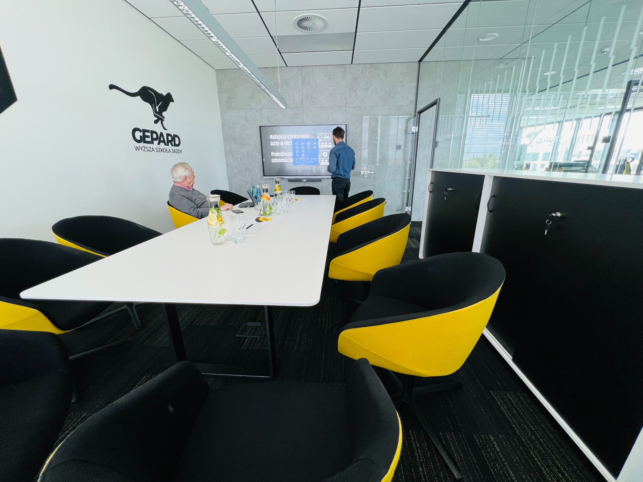 Sala konferencyjna z białym stołem, czarno-żółtymi krzesłami i logiem Gepard Wyższa Szkoła Jazdy na ścianie. Mężczyzna stoi przy interaktywnej tablicy z napisem 'Najlepsza szkoła nauki jazdy...