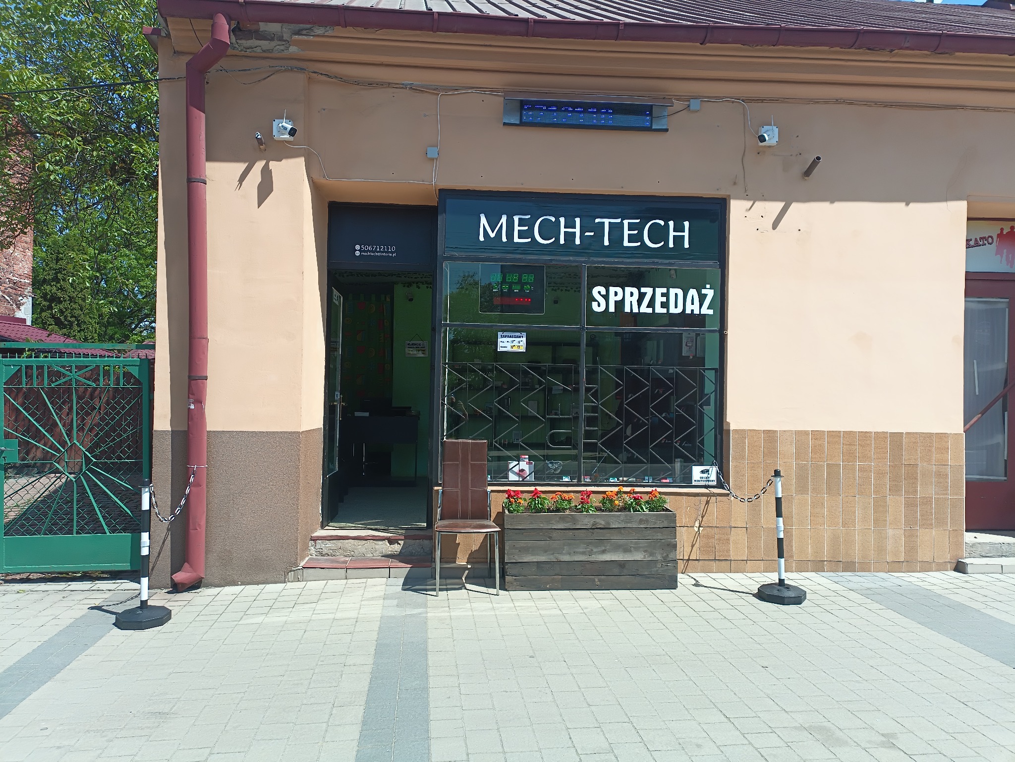 Fasada budynku firmy Mech-Tech z widocznymi kamerami monitoringu, okno wystawowe z napisem 'Sprzedaż' i wyświetlaczem LED, wejście z krzesłem i skrzynią z kwiatami.