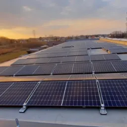 Rząd paneli słonecznych na płaskim dachu budynku przemysłowego w promieniach zachodzącego słońca, z widoczną linią energetyczną i zabudowaniami w tle.