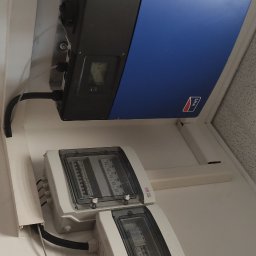 Instalacje Elektryczne Artur Okniński - Niebiesko-czarny inwerter marki SMA zamontowany na ścianie obok dwóch białych rozdzielnic elektrycznych z przezroczystymi pokrywami.