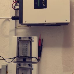 Instalacje Elektryczne Artur Okniński - Biały inwerter fotowoltaiczny marki FOX ESS zamontowany na ścianie, poniżej dwie skrzynki z bezpiecznikami, widoczne przewody i mierniki elektryczne, szara rura instalacyjna na górze.