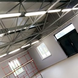 Instalacje Elektryczne Artur Okniński - Wnętrze hali w Łukowie z metalową konstrukcją dachu, oświetleniem LED, rusztowaniem i oknami. Ściany szare, podłoga jasna.