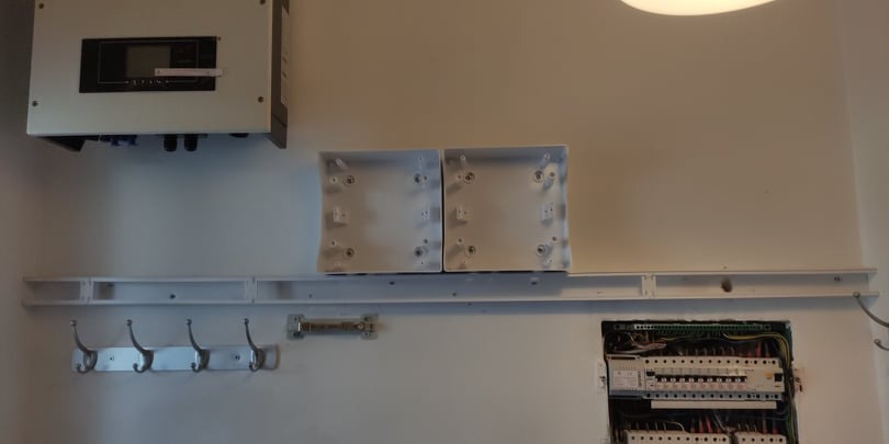 Wnętrze pomieszczenia z zamontowaną skrzynką elektryczną, inwerterem fotowoltaicznym i rozdzielnicą elektryczną z widocznymi przewodami, poniżej wieszak na ubrania z haczykami.