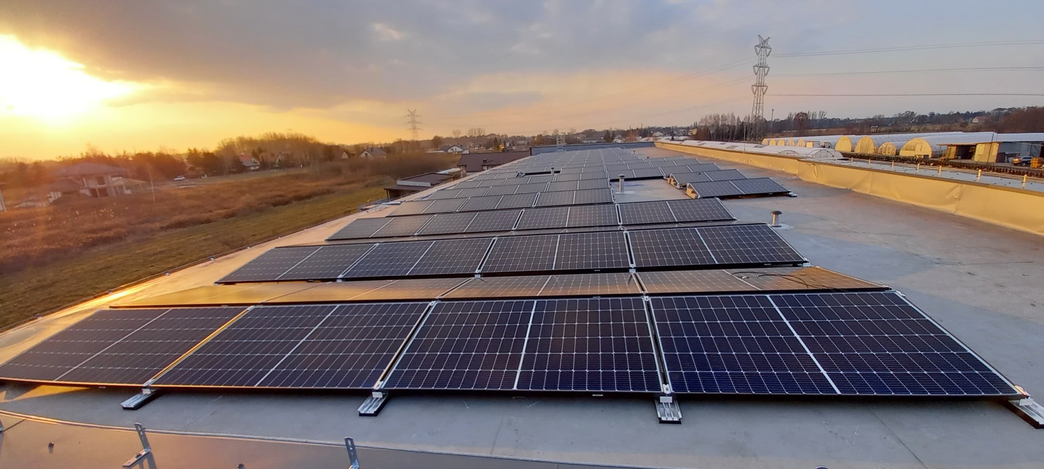 Rząd paneli słonecznych na płaskim dachu budynku przemysłowego w promieniach zachodzącego słońca, z widoczną linią energetyczną i zabudowaniami w tle.