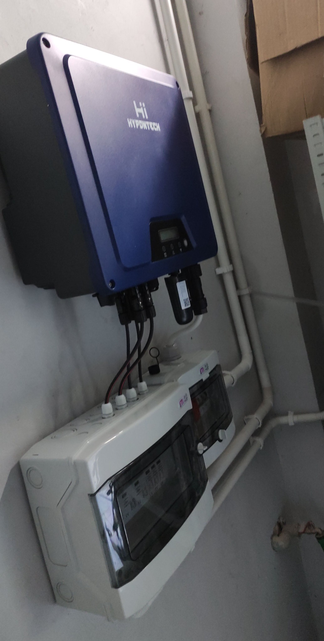 Niebieski inwerter Hypontech zamontowany nad białą skrzynką rozdzielczą ETI na szarej ścianie, widoczne przewody i rurki instalacyjne.