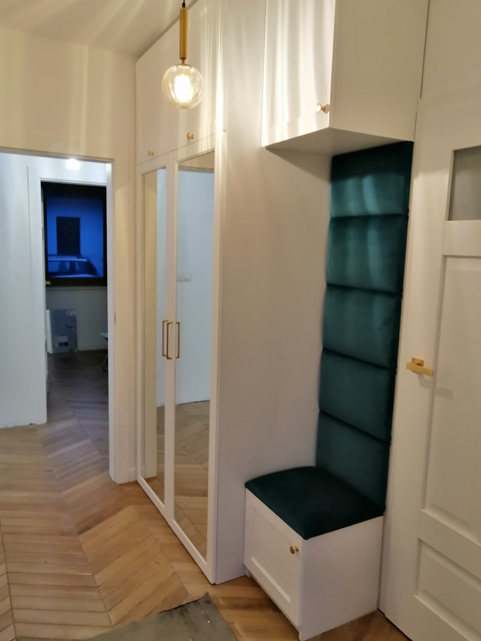 Biała szafa z lustrzanymi drzwiami i siedziskiem z zielonym tapicerowanym panelem ściennym, oświetlona wiszącą lampą z kulistym kloszem, parkiet w jodełkę na podłodze.