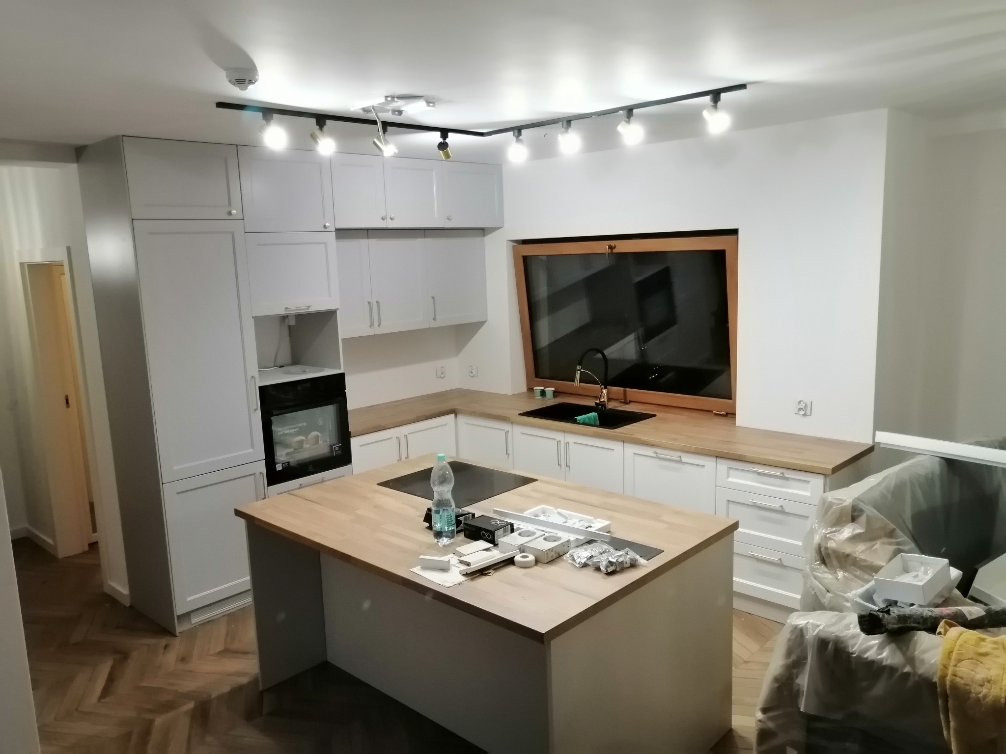 Jasna kuchnia z drewnianym blatem i wyspą, lampy sufitowe szynowe, sprzęty i akcesoria na wyspie, podłoga w jodełkę, okno w drewnianej ramie nad zlewem.