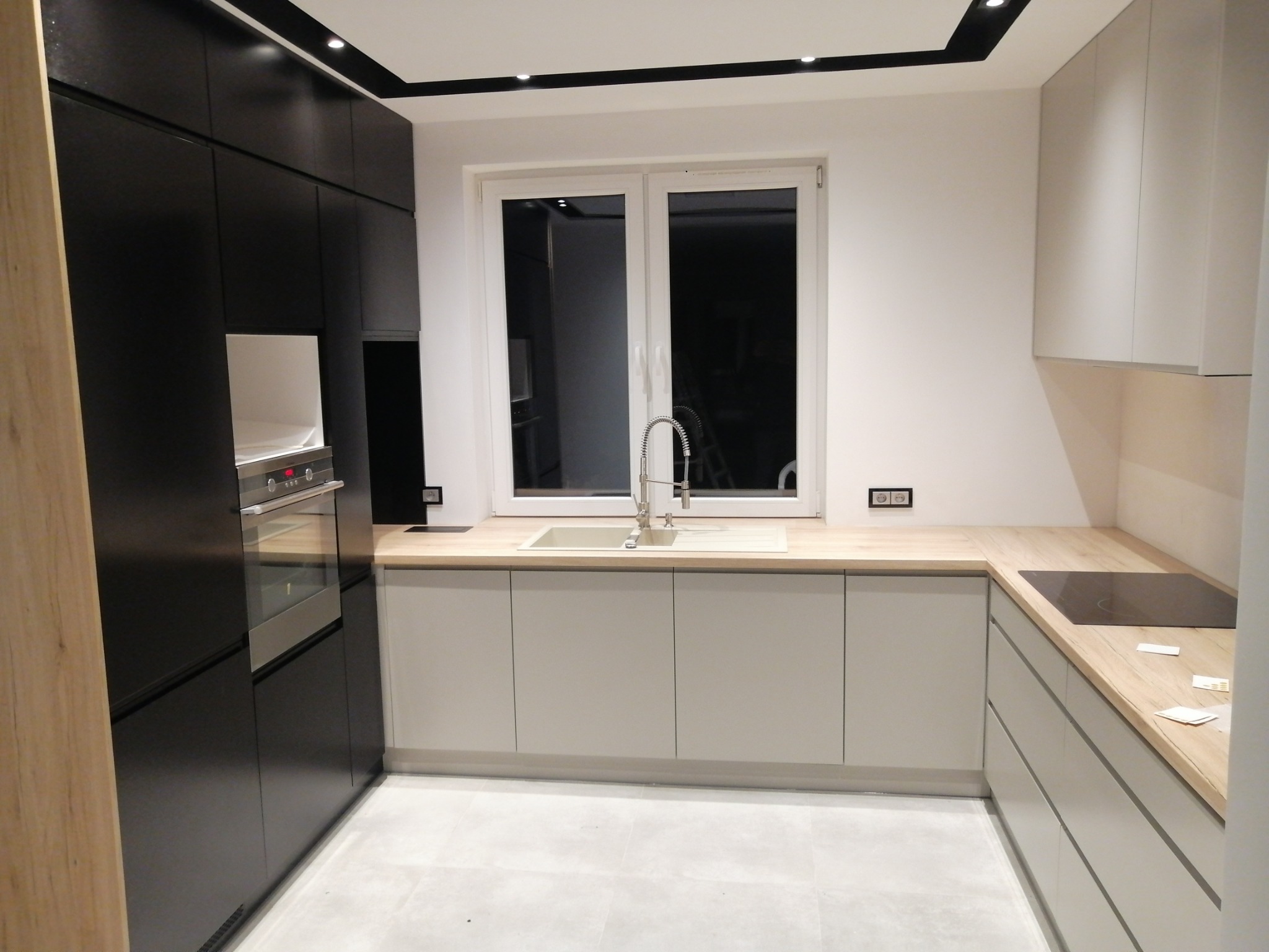 Nowoczesna kuchnia z czarnymi szafkami, piekarnikiem w zabudowie, jasnymi szafkami dolnymi i drewnianym blatem, widok na zlewozmywak i płytę indukcyjną.