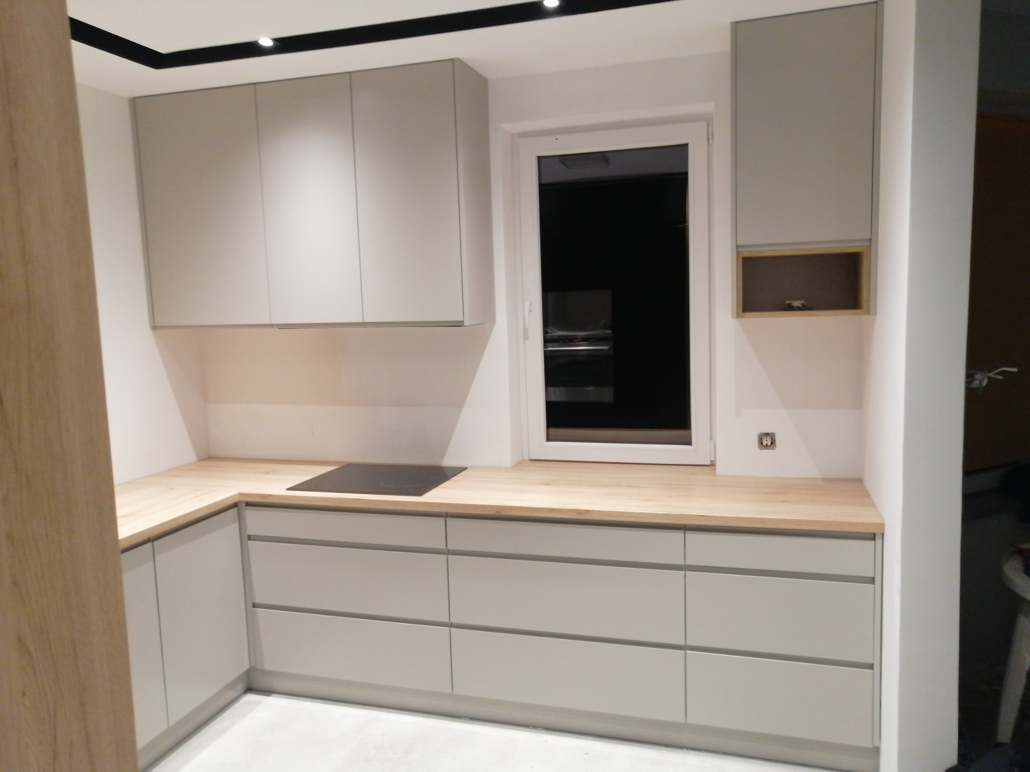 Jasnoszara zabudowa kuchenna z drewnianym blatem, płyta indukcyjna wpuszczona w blat, okno w tle, minimalistyczny styl.