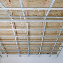 Sławomir Kuczkowicz Termowizja i termoizolacja - Metalowa konstrukcja sufitowa z wypełnieniem izolacyjnym, widok z dołu, białe ściany w tle.