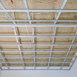 Metalowa konstrukcja sufitowa z wypełnieniem izolacyjnym, widok z dołu, białe ściany w tle.