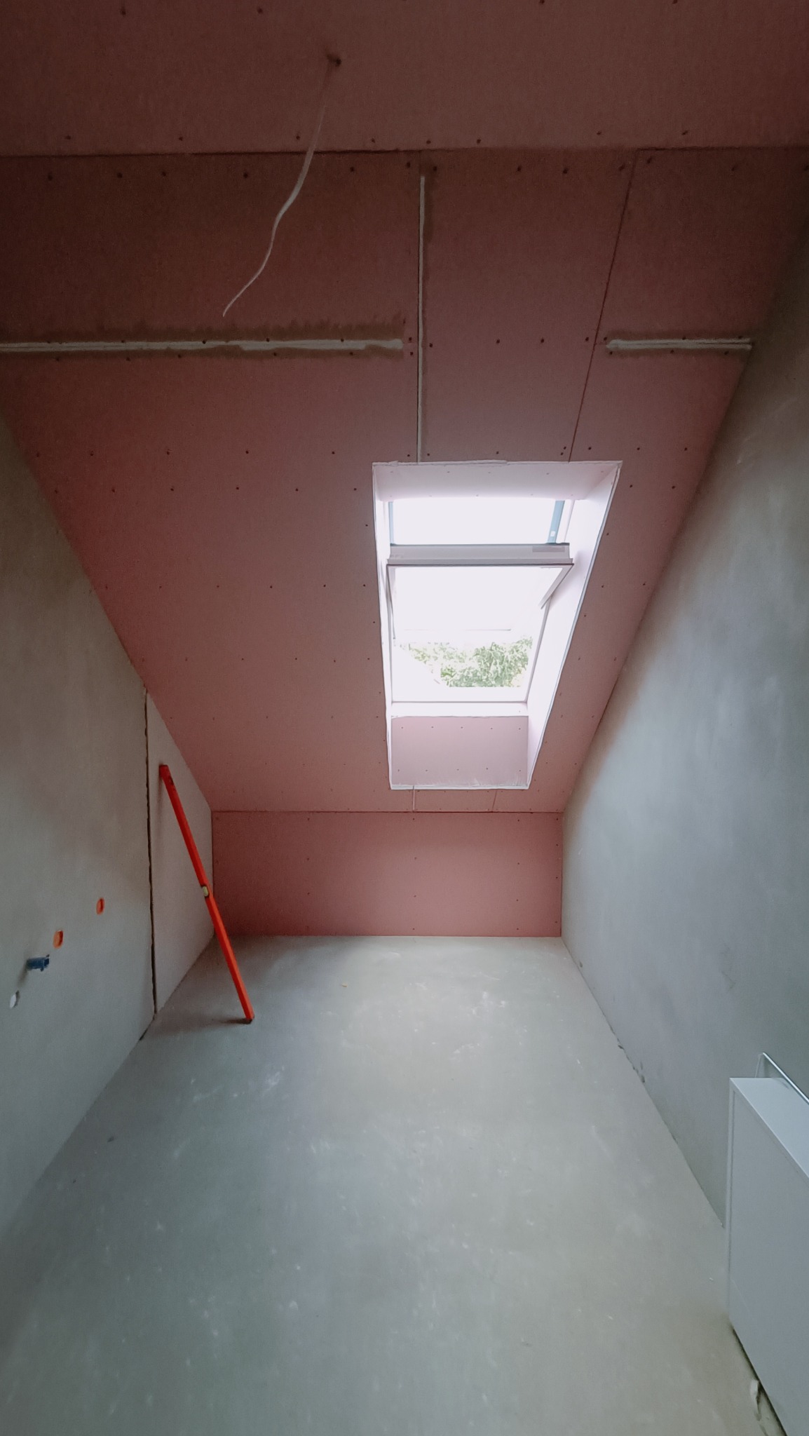 Poddasze w trakcie adaptacji: widoczne różowe płyty na skosach, okno dachowe wpuszczające światło, surowa podłoga i poziomica oparta o ścianę.