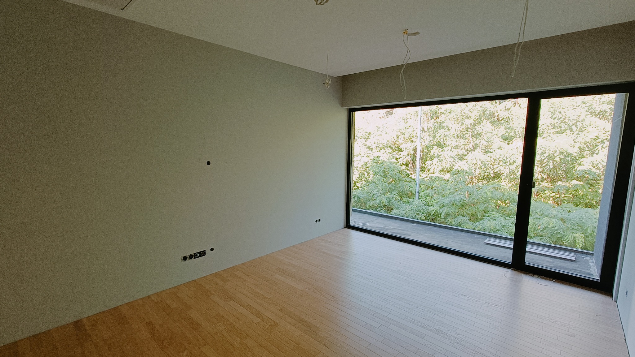 Wnętrze pokoju z parkietem, gładkimi ścianami w odcieniach szarości, dużym oknem z widokiem na zieleń, widoczne gniazdka elektryczne i wyprowadzone kable oświetleniowe.