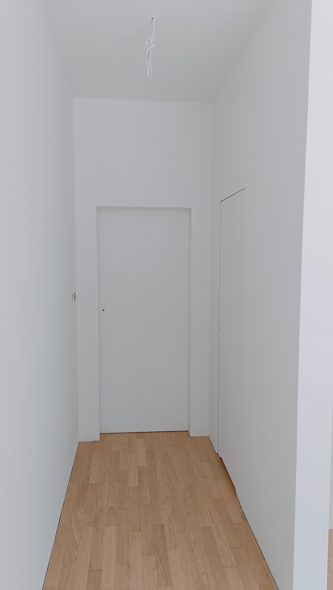 Wnętrze korytarza po remoncie: świeżo pomalowane białe ściany, jasna drewniana podłoga, minimalistyczne drzwi w kolorze ścian, widoczny przewód oświetleniowy na suficie.