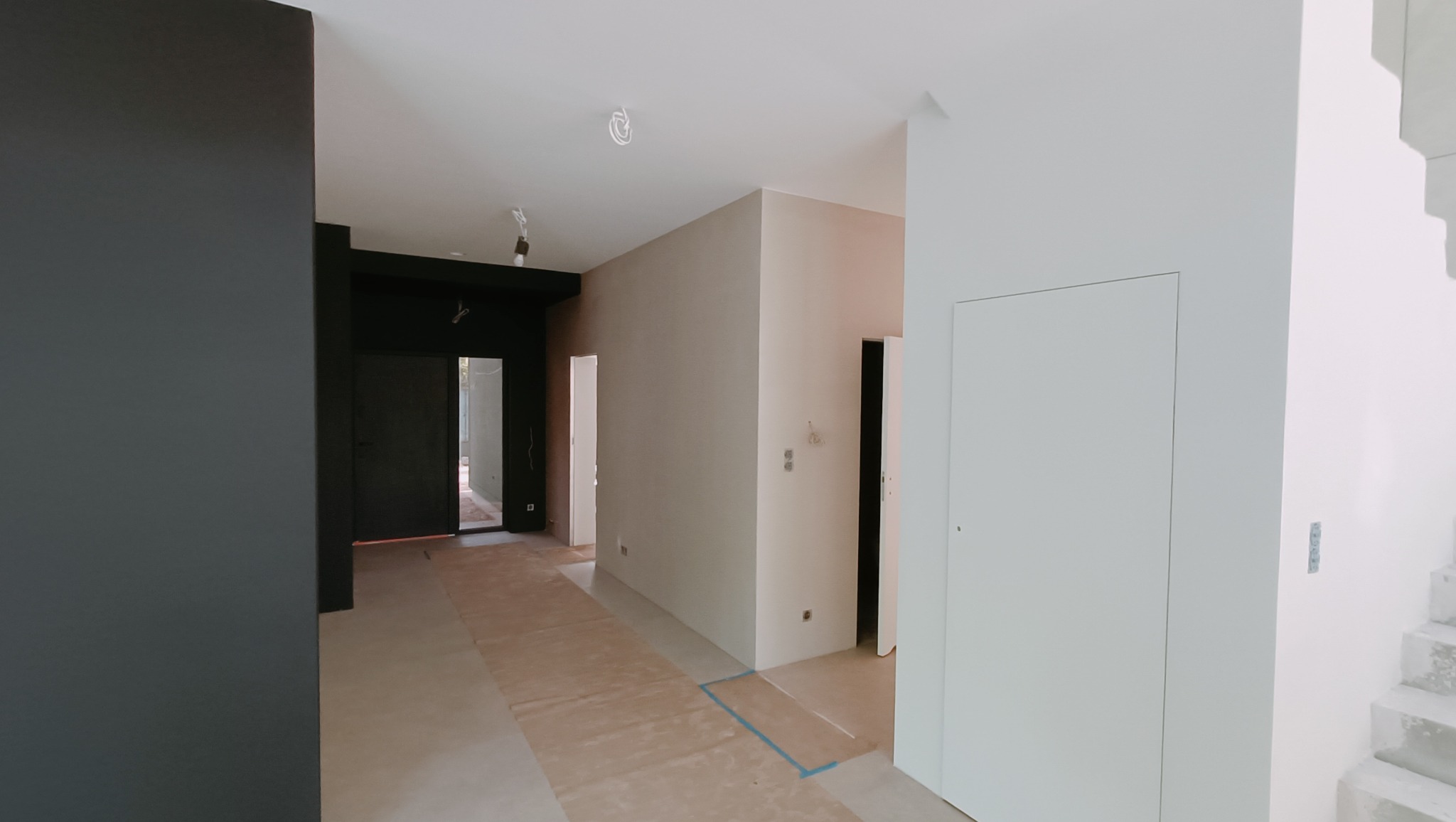 Wnętrze w trakcie wykańczania: zabezpieczona podłoga, pomalowane ściany na jasny beż i ciemny grafit, widoczne schody i ukryte drzwi.