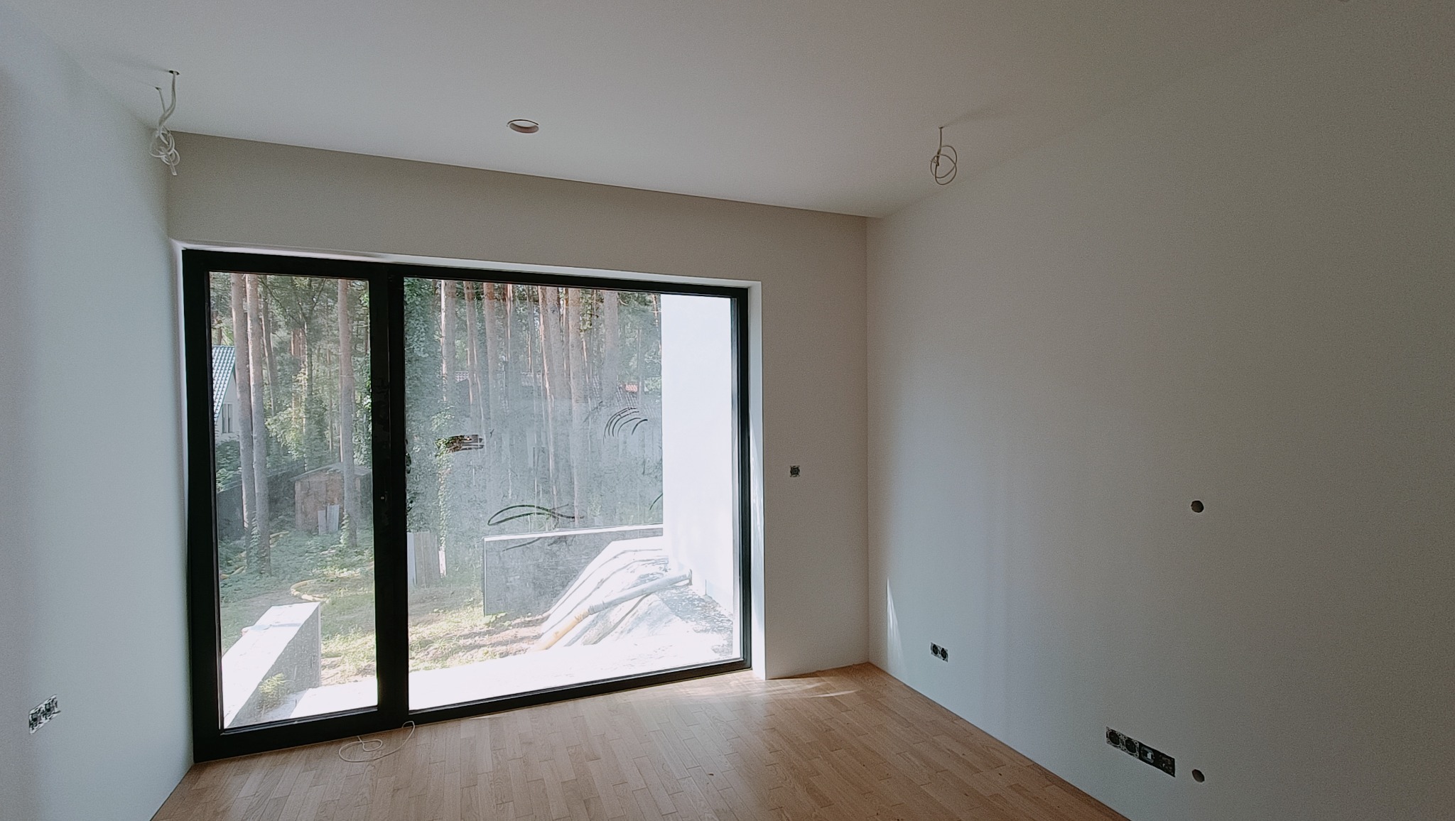 Surowe wnętrze z drewnianą podłogą, dużym oknem z widokiem na las, niedokończonymi instalacjami elektrycznymi w ścianach i na suficie.