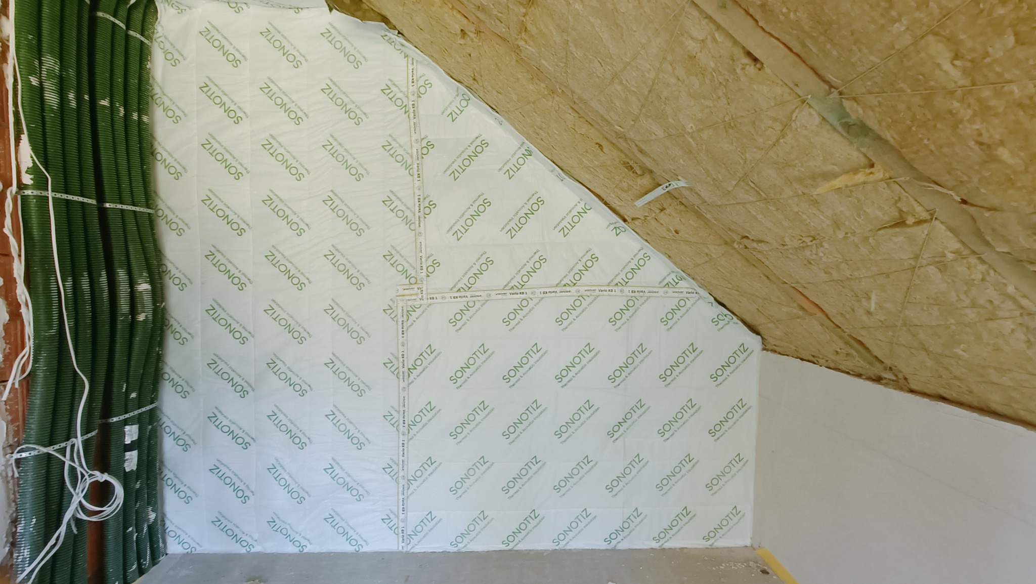 Poddasze w trakcie ocieplania: widoczna wełna mineralna oraz folia paroizolacyjna z nadrukiem 'SONOTIZ' i zielone rury wentylacyjne przy ścianie.