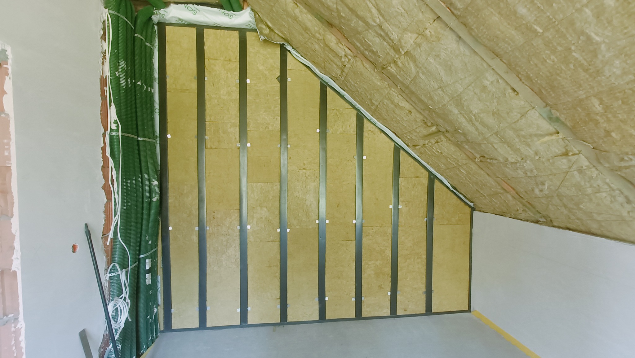 Poddasze w trakcie ocieplania: widoczne metalowe profile konstrukcyjne, żółta wełna mineralna między profilami oraz częściowo ułożona folia paroizolacyjna na skośnym dachu, z boku ściana z rurami...