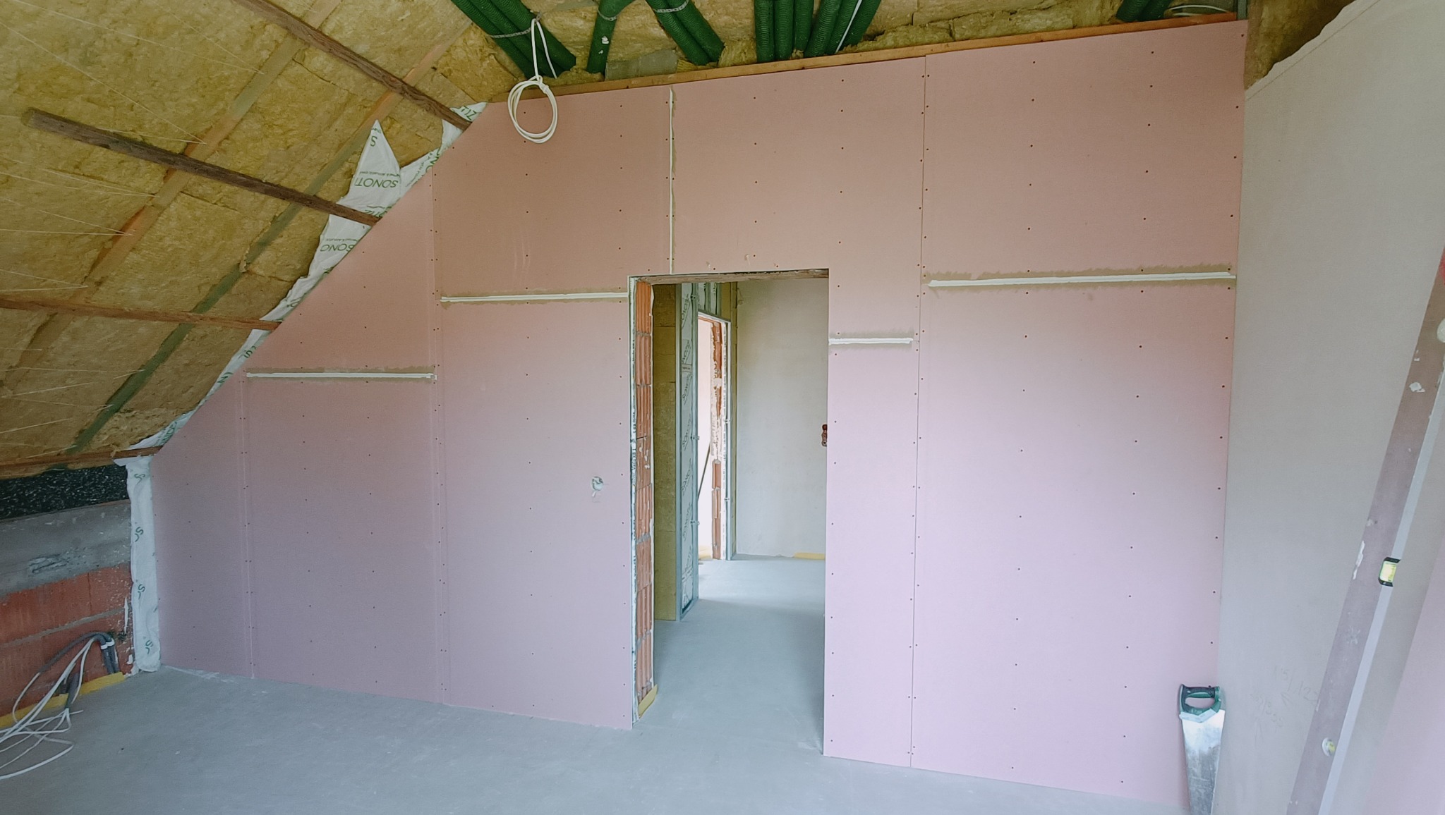 Poddasze z ociepleniem i zamontowanymi płytami w kolorze różowym, widoczne wełna mineralna, profile i instalacje pod sufitem oraz otwór drzwiowy do kolejnego pomieszczenia.