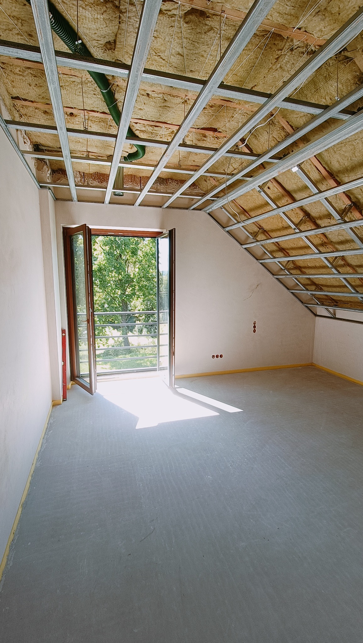 Poddasze w trakcie ocieplania, widoczne wełna mineralna i metalowy stelaż sufitowy, otwarte drzwi balkonowe wpuszczają światło słoneczne.