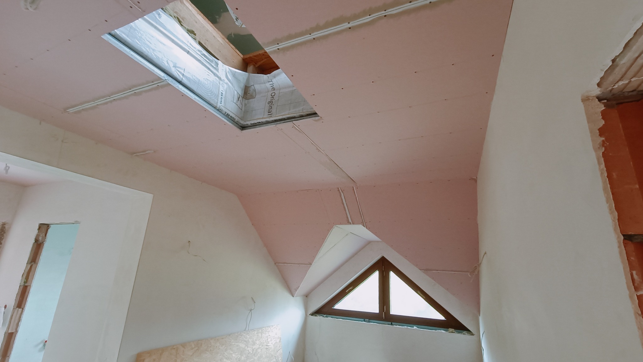 Montaż sufitu podwieszanego z płyt gipsowo-kartonowych na poddaszu, widoczne okno dachowe i trójkątne okno w ścianie szczytowej, ściany przygotowane do malowania.