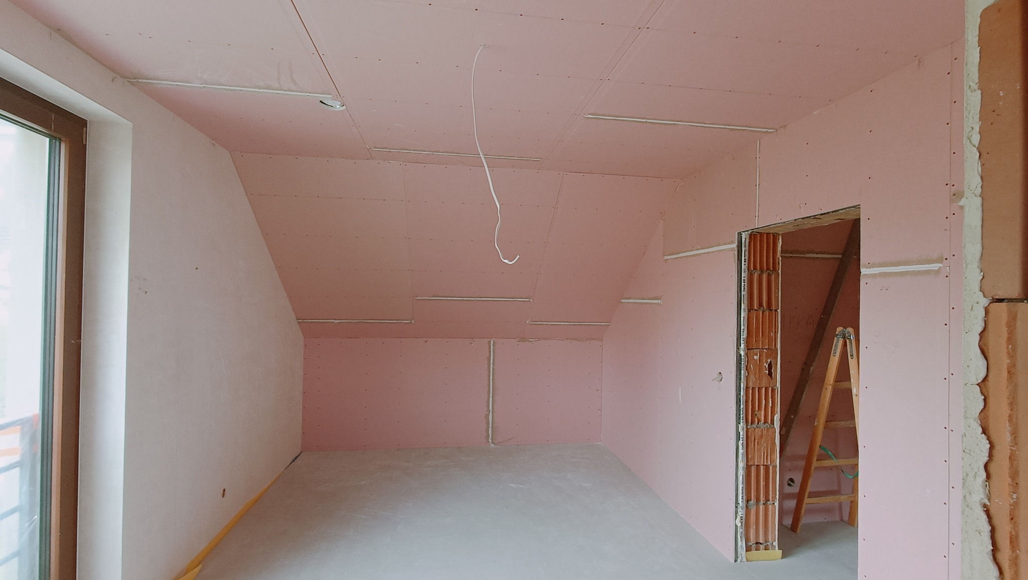 Poddasze z zamontowanymi płytami w kolorze różowym, widoczne profile i przewody elektryczne, w tle drabina oparta o ścianę.
