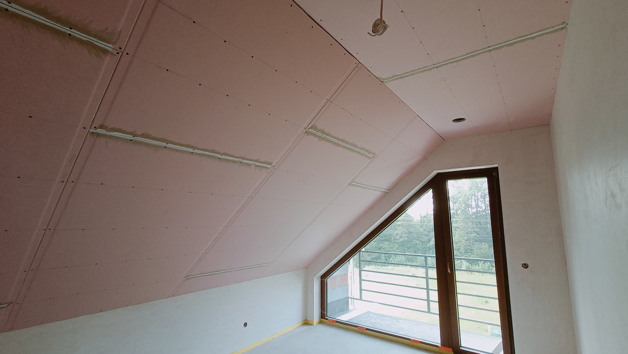 Poddasze w trakcie wykańczania: widoczne płyty gipsowo-kartonowe na skosach, drewniane okno trójkątne z widokiem na zieleń, surowa ściana i podłoga.