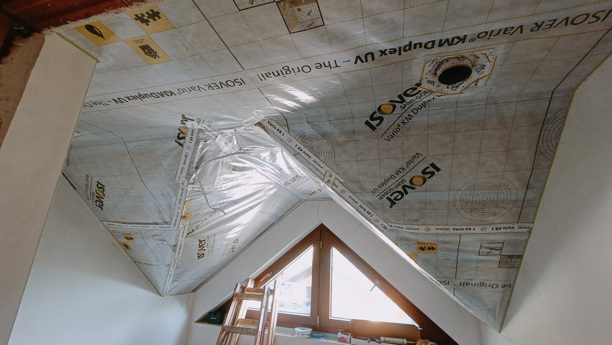 Poddasze w trakcie ocieplania, widoczna folia paroizolacyjna Isover Vario KM Duplex UV, okno dachowe i drabina oparta o ścianę.