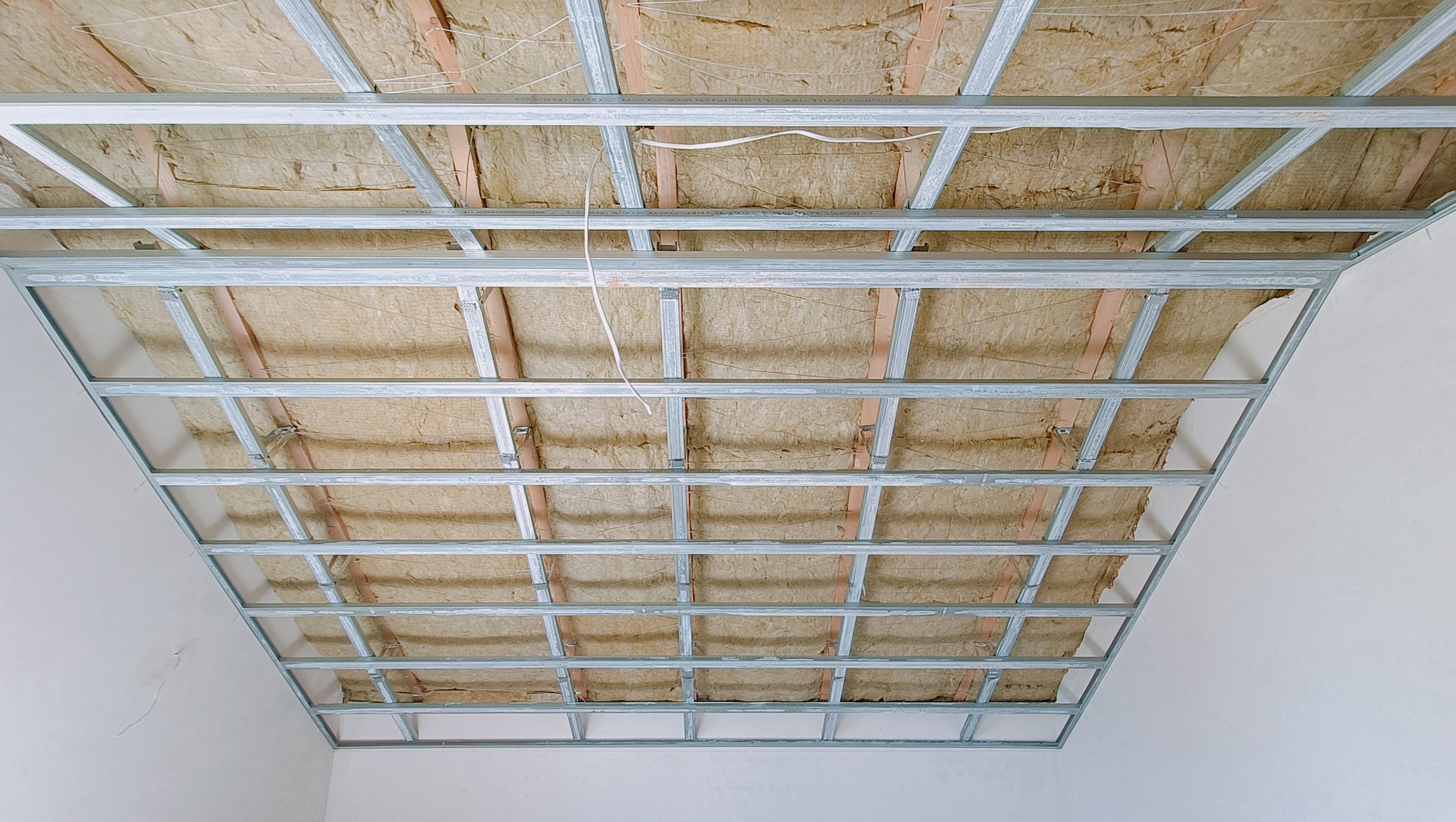 Metalowa konstrukcja sufitowa z wypełnieniem izolacyjnym, widok z dołu, białe ściany w tle.