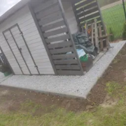 Drewniana szopa z szarymi elementami, otoczona obrzeżem i wysypana białym grysem, widoczne narzędzia ogrodnicze i palety