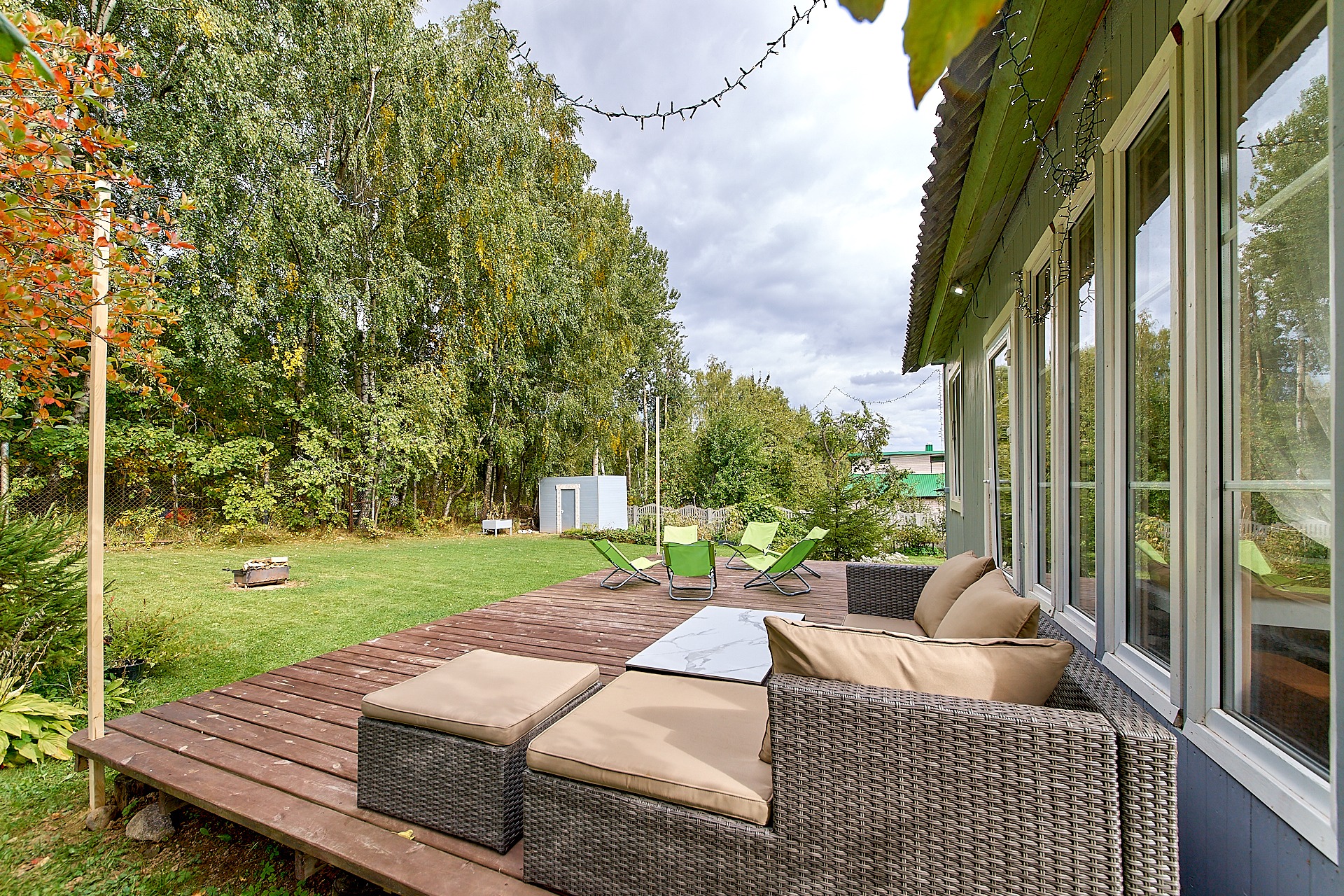 Rustykalny taras z ciemnego drewna z rattanowym zestawem wypoczynkowym w odcieniach beżu, na trawniku zielone leżaki i palenisko, w tle drzewa i mały domek gospodarczy.
