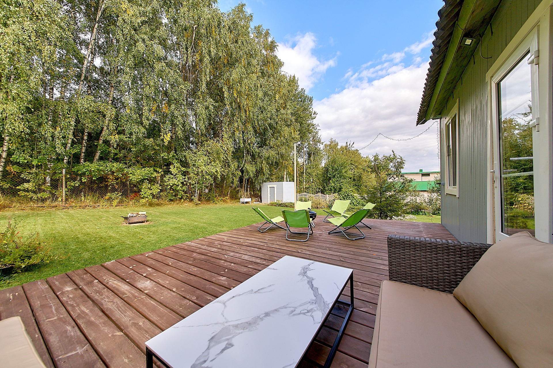 Drewniany taras z meblami ogrodowymi: sofa z wikliny, marmurowy stolik kawowy i zielone leżaki, widok na trawnik z grillem i drzewa w tle.