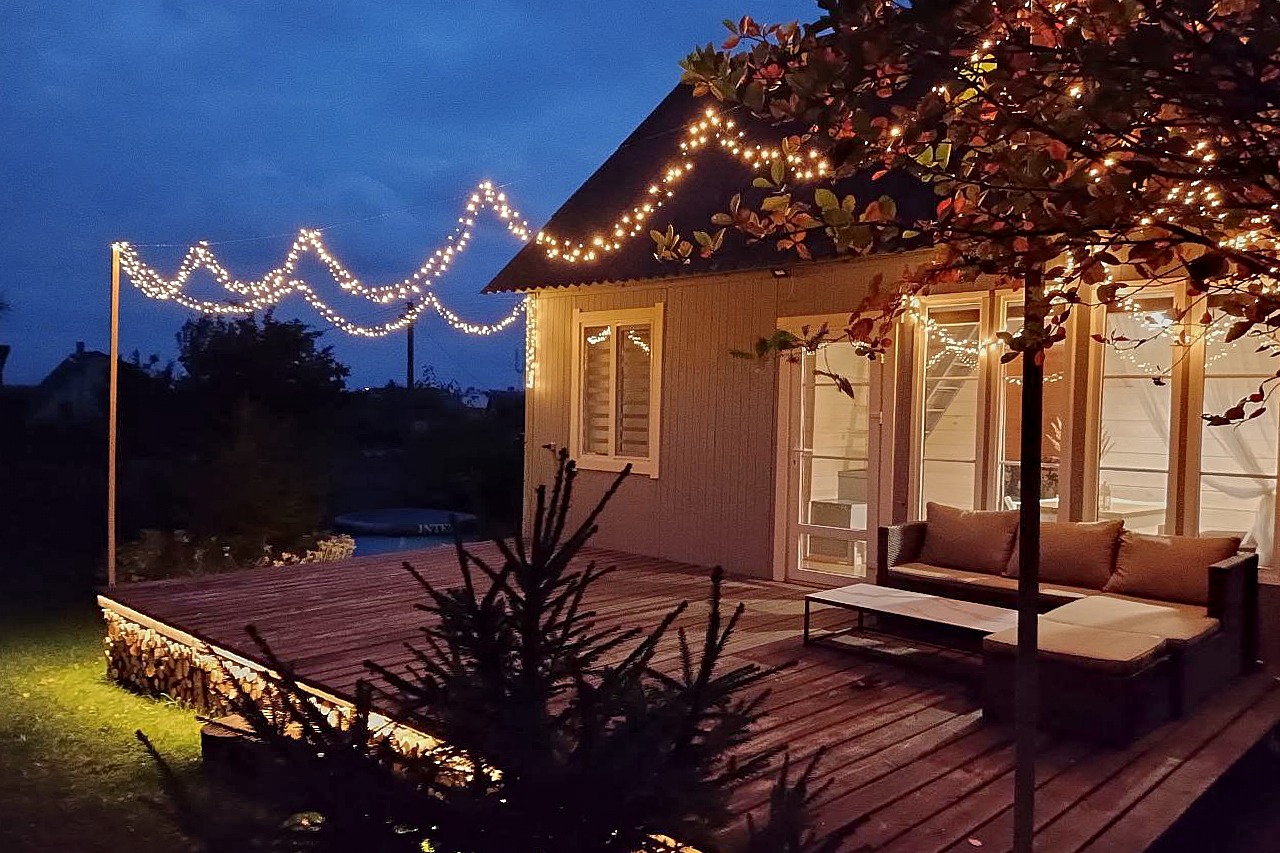 Wieczorny widok na drewniany taras z oświetleniem girlandami, szary dom z dużymi oknami, meble ogrodowe i drzewo z liśćmi w jesiennych barwach.