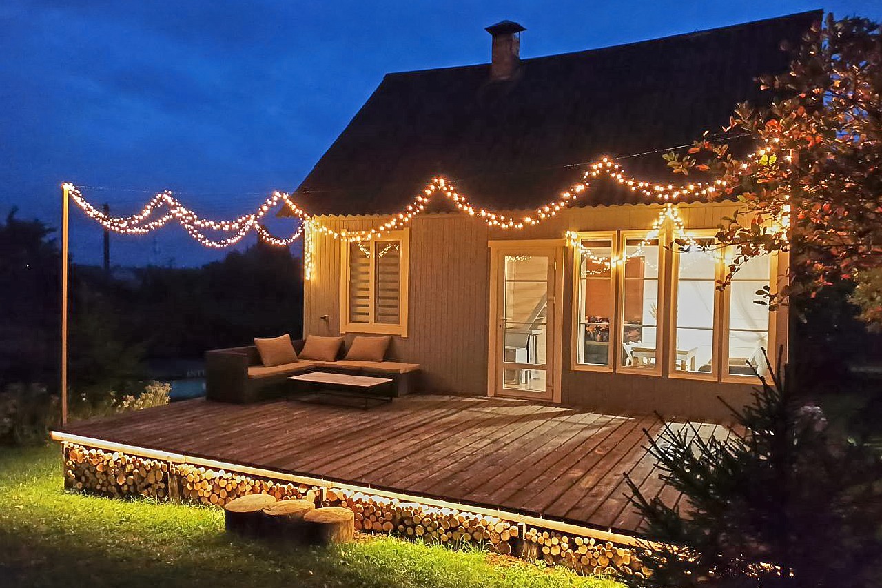 Oświetlony drewniany taras z meblami wypoczynkowymi i dekoracyjnymi lampkami sznurkowymi, obłożony drewnem opałowym, widok na dom o zmierzchu.