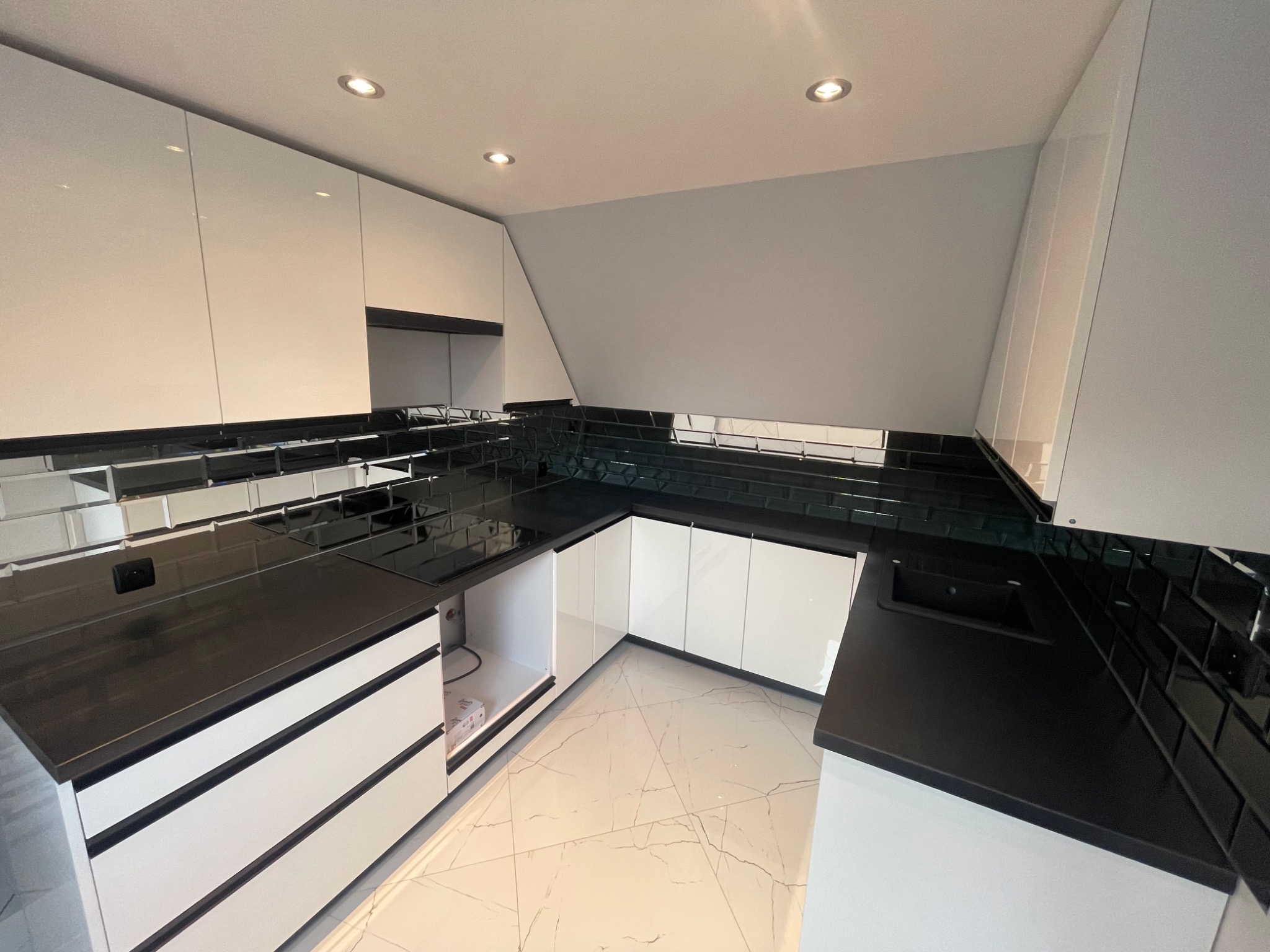 Nowoczesna kuchnia w czerni i bieli: błyszczące, białe szafki z czarnymi blatami i mozaiką, marmurowa podłoga, oświetlenie punktowe.